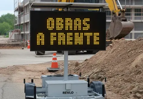 Painel de mensagem variável móvel exibindo OBRAS À FRENTE em canteiro com escavadeira